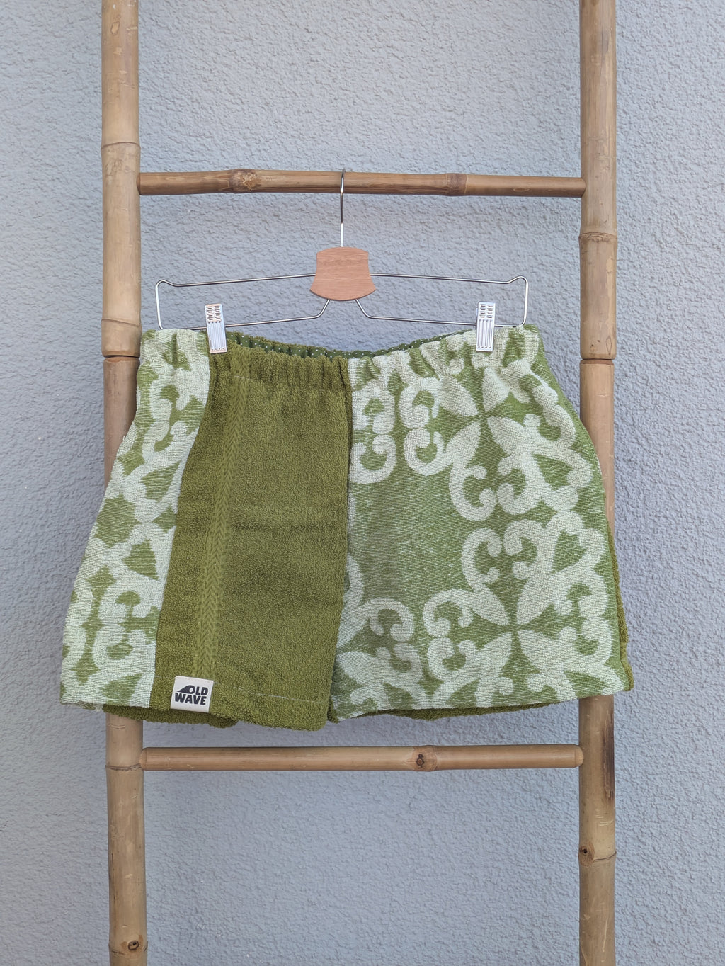 Vorderansicht der Frottee Towel Shorts 'Olive Ornament ’75' von Old Wave in Olivgrün und Salbeigrün. Patchwork-Design mit großflächigem barocken Fliesenmuster, unifarbenem grünen Hosenbeineinsatz und Old Wave Logo-Patch unten links, präsentiert an einer Bambusleiter.