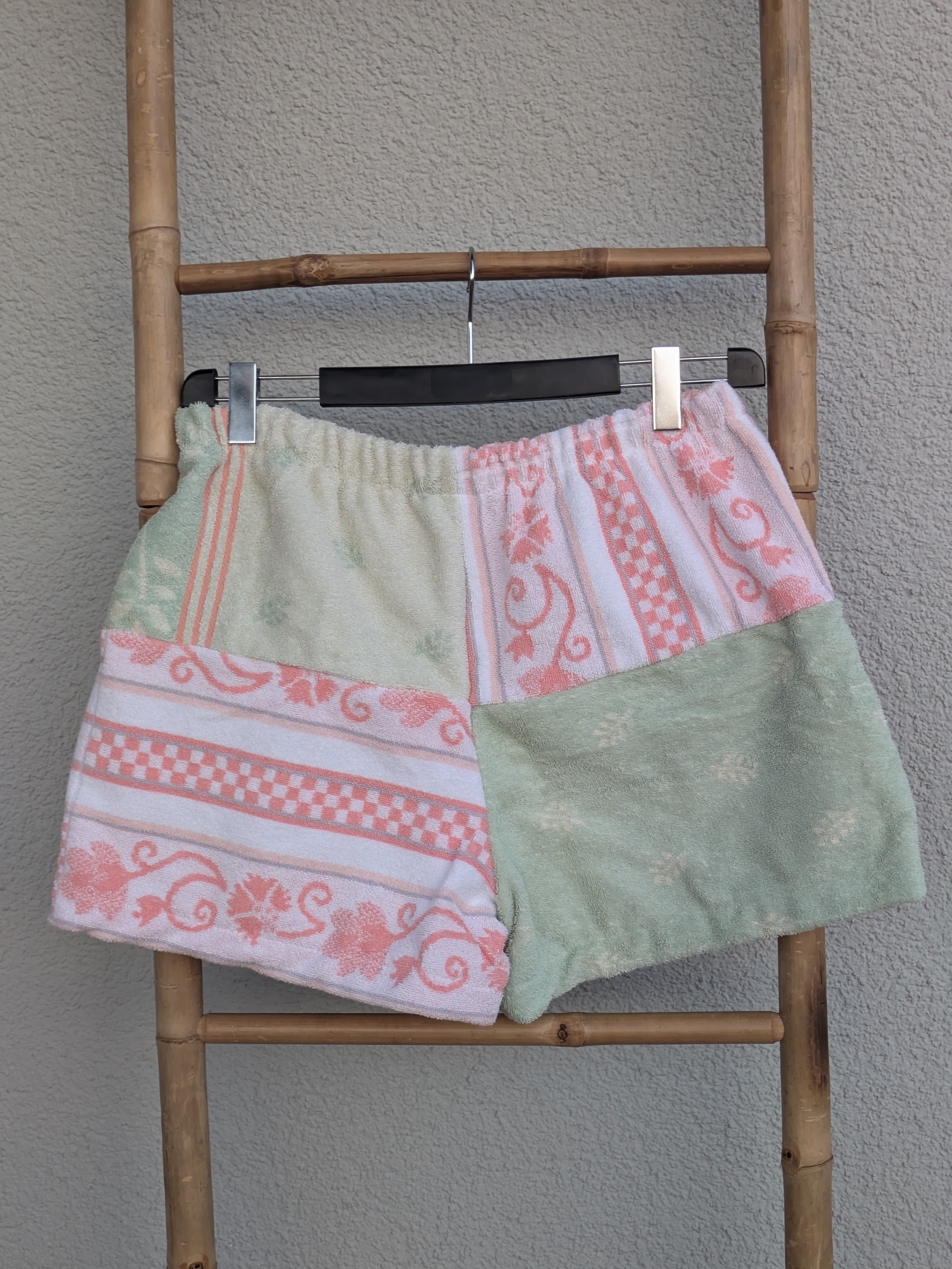 Rückansicht der Old Wave Towel Shorts 'Peach Mint Mosaic ’73'. Überwiegend mintgrünes Frottee mit hellen Mustereinsätzen und feinem Pfirsich-Akzentstreifen. Die Shorts hängen an einer Bambusleiter.