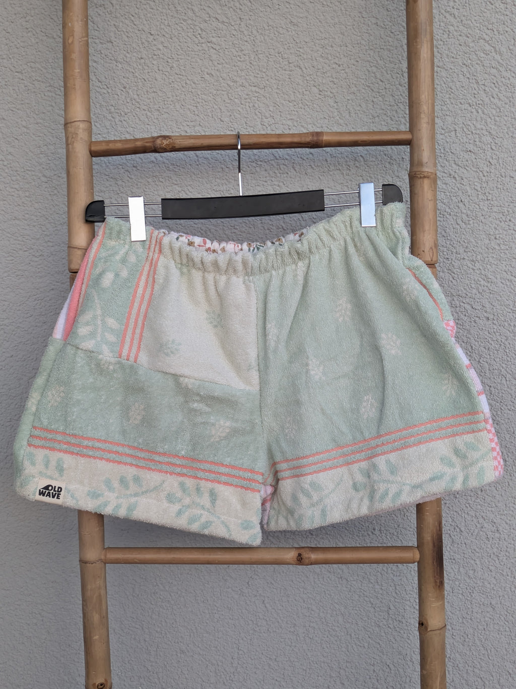 Vorderansicht der Frottee-Shorts 'Peach Mint Mosaic ’73' von Old Wave. Zeigt ein Patchwork-Design aus mintgrünem Stoff und pfirsichfarben-weißen Retro-Musterborden. Die Hose hängt an einem Kleiderbügel vor einer hellen Wand.