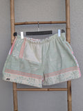 Vorderansicht der Frottee-Shorts 'Peach Mint Mosaic ’73' von Old Wave. Zeigt ein Patchwork-Design aus mintgrünem Stoff und pfirsichfarben-weißen Retro-Musterborden. Die Hose hängt an einem Kleiderbügel vor einer hellen Wand.