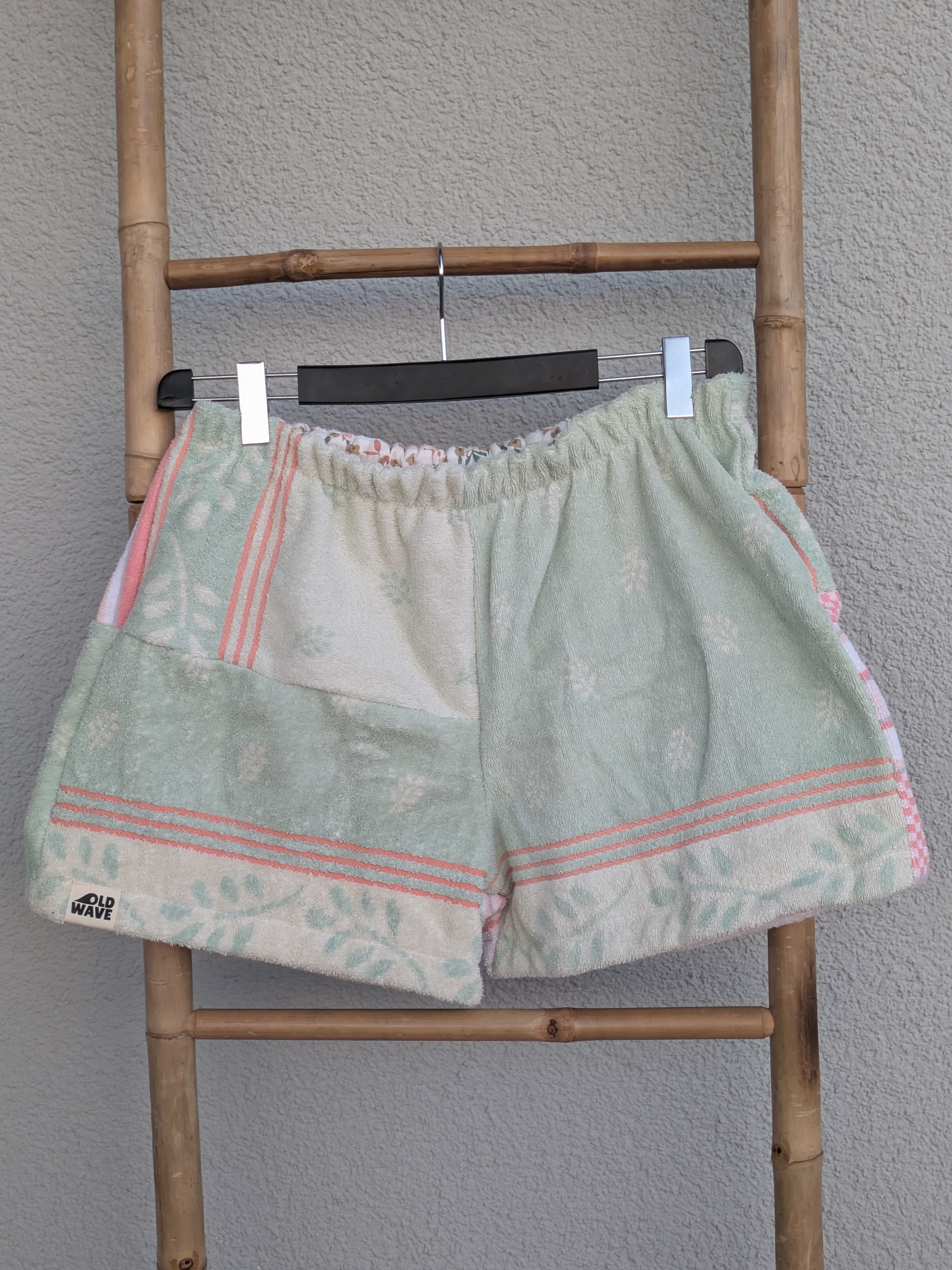 Vorderansicht der Frottee-Shorts 'Peach Mint Mosaic ’73' von Old Wave. Zeigt ein Patchwork-Design aus mintgrünem Stoff und pfirsichfarben-weißen Retro-Musterborden. Die Hose hängt an einem Kleiderbügel vor einer hellen Wand.