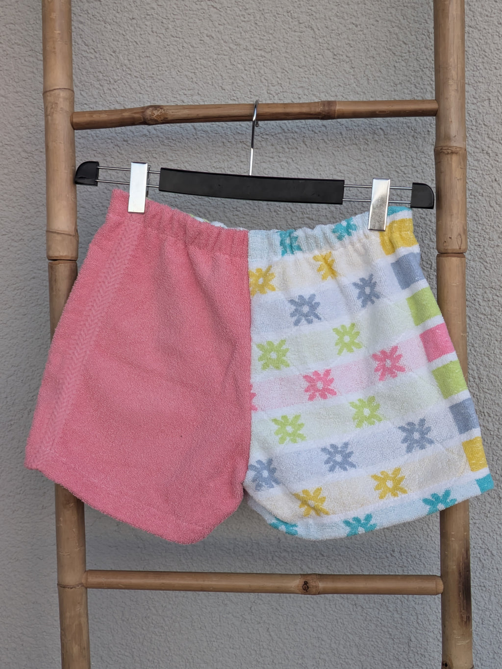 Rückansicht der Frottee-Shorts 'Pixel Punch ’78' von Old Wave im Half-and-Half-Design: Das linke Hosenbein ist einfarbig altrosa, das rechte Hosenbein zeigt ein buntes geometrisches Muster. Die Hose hängt an einer Bambusleiter.