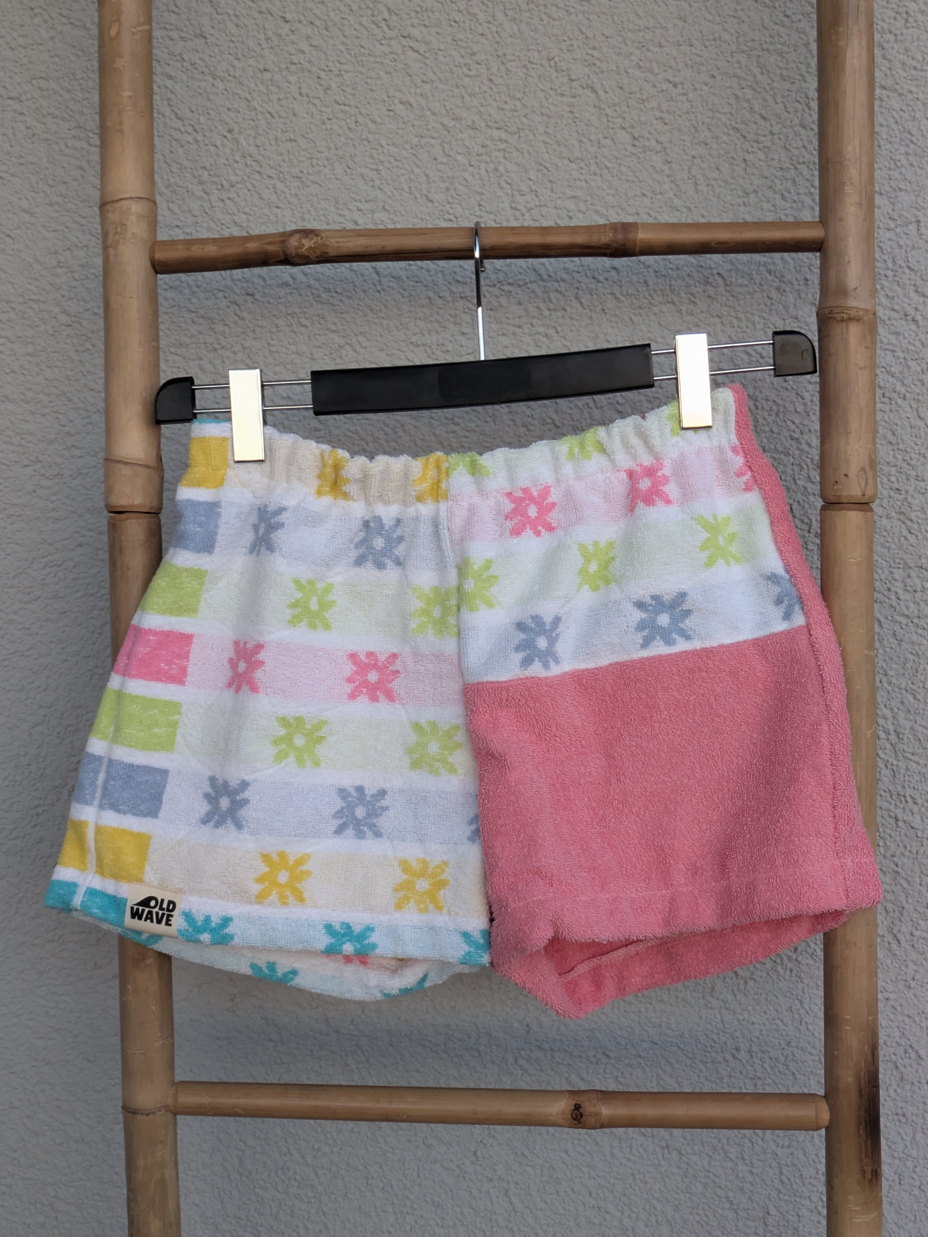 Vorderansicht der Old Wave Towel Shorts 'Pixel Punch ’78'. Buntes Retro-Kästchenmuster kombiniert mit einem altrosafarbenen Hosenbein und gemustertem Bund. Die Shorts hängen an einem Kleiderbügel vor einer hellen Wand.