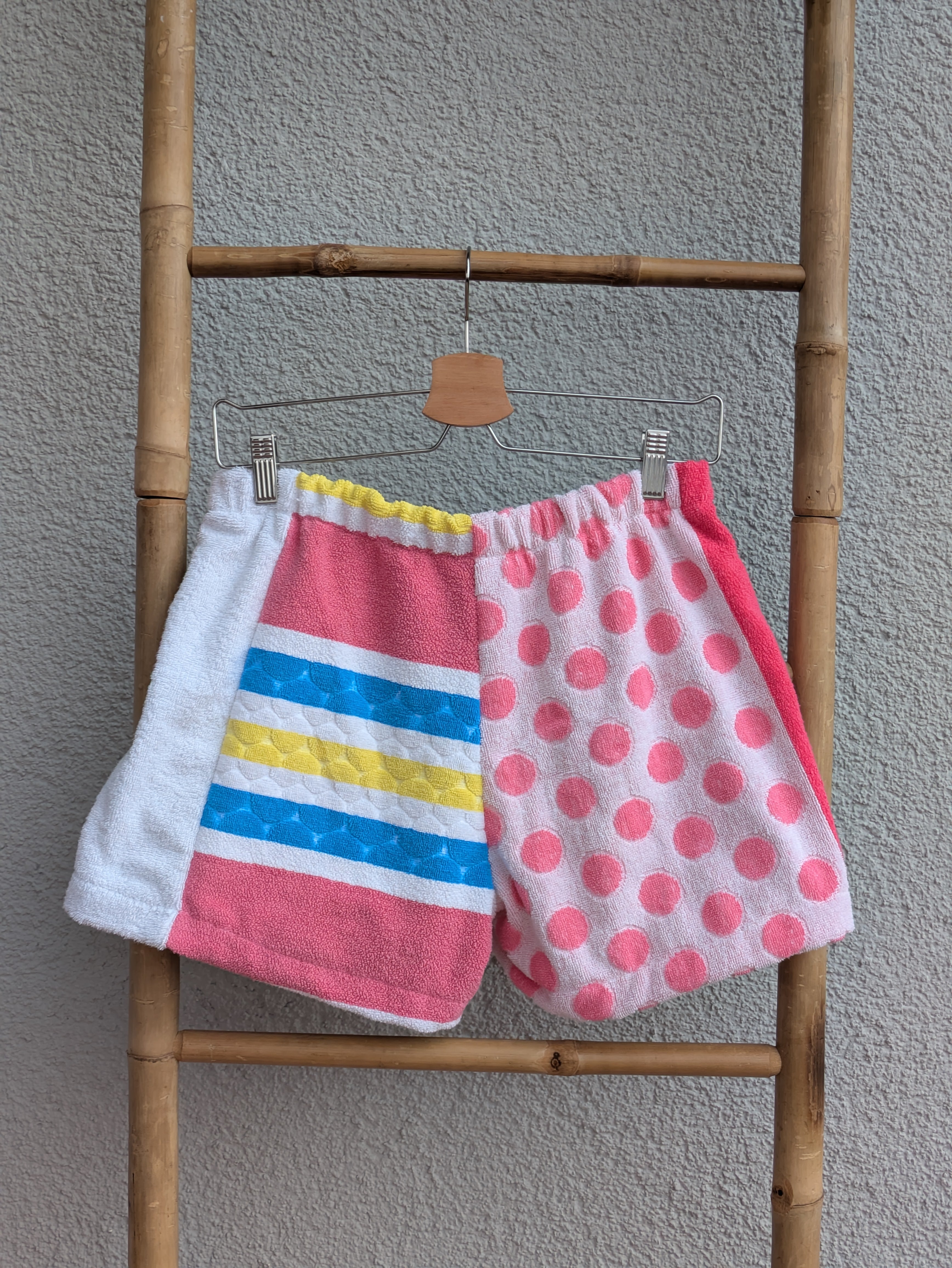 Rückansicht der Frottee Towel Shorts 'Retro Festival Dots ’76' von Old Wave. Zeigt das verspielte Patchwork-Design mit quer gestreiftem Block in Pink, Blau und Gelb links sowie rosa Polka Dots auf der rechten Hosenhälfte vor einer hellen Wand.