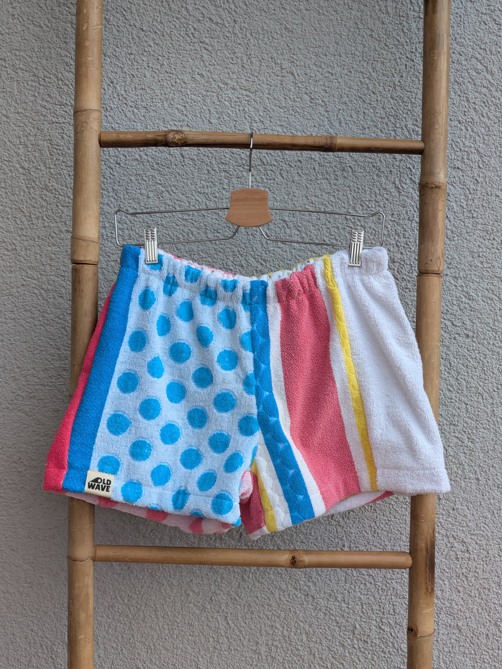 Vorderansicht der Frottee Towel Shorts 'Retro Festival Dots ’76' von Old Wave. Bunter Patchwork-Mix mit blauem Polka-Dot-Muster links, pinken und gelben Längsstreifen sowie weißem Frottee-Hosenbein rechts, präsentiert an einer Bambusleiter.