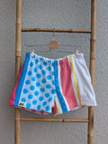 Vorderansicht der Frottee Towel Shorts 'Retro Festival Dots ’76' von Old Wave. Bunter Patchwork-Mix mit blauem Polka-Dot-Muster links, pinken und gelben Längsstreifen sowie weißem Frottee-Hosenbein rechts, präsentiert an einer Bambusleiter.