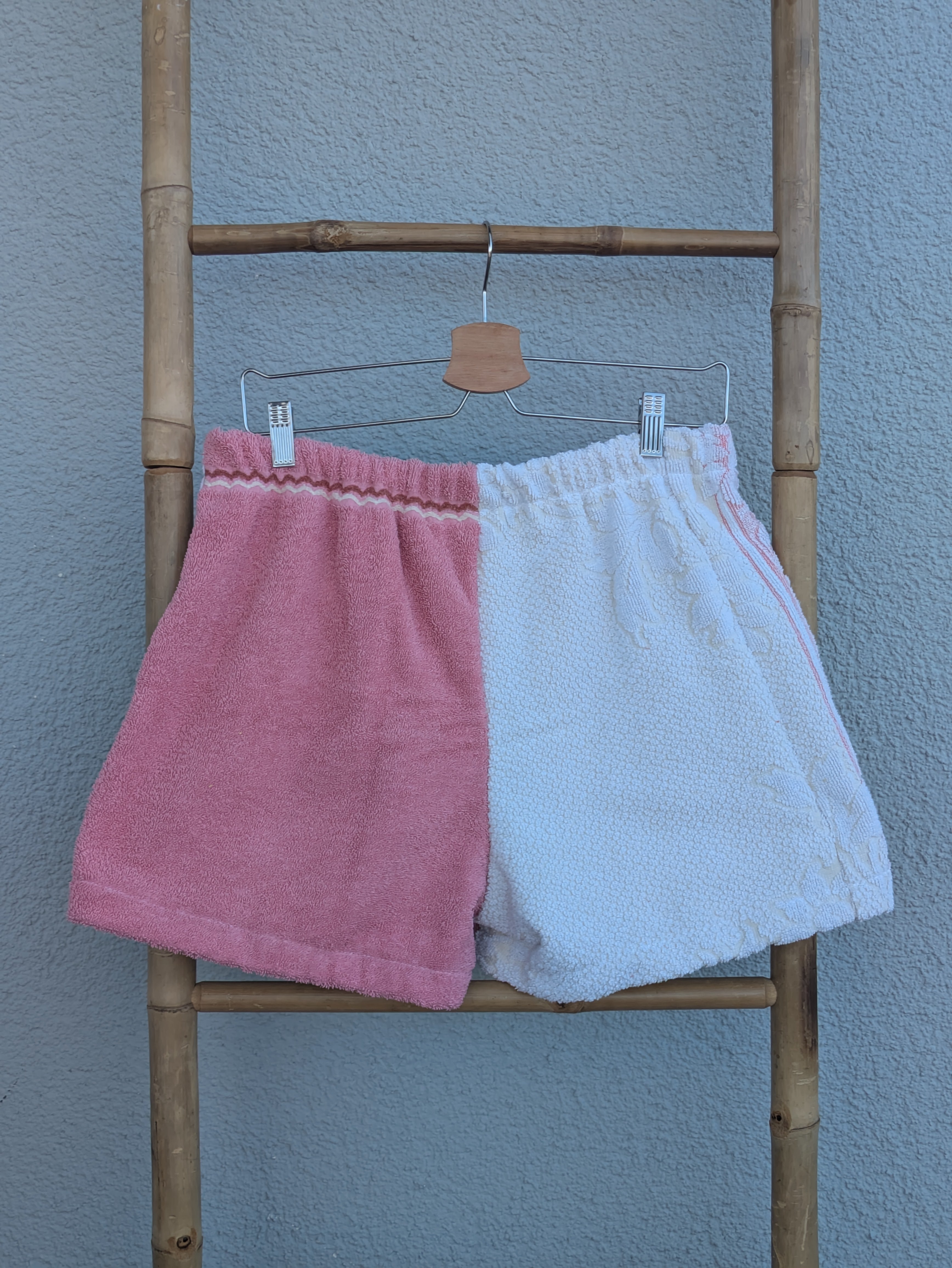 Rückansicht der Frottee Towel Shorts 'Retro Rose Patch ’79' von Old Wave. Zeigt das zweigeteilte Half-and-Half-Design in unifarbenem Altrosa und hellem Frotteestoff vor einer hellen Wand.