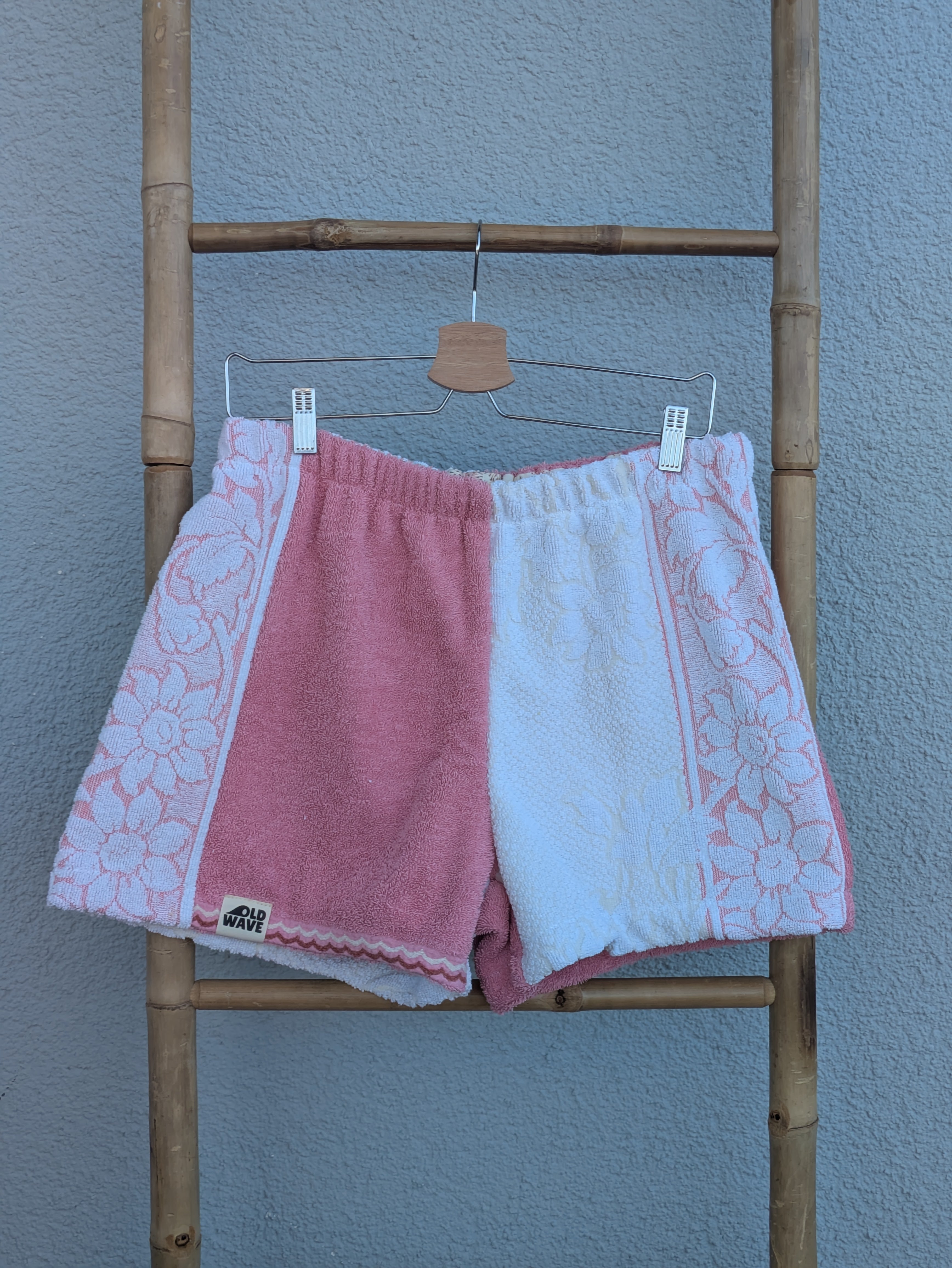 Vorderansicht der Frottee Towel Shorts 'Retro Rose Patch ’79' von Old Wave in Altrosa, Weiß und Zartrosa. Mehrfarbiges Patchwork-Design mit gemustertem Seiteneinsatz, unifarbener Mitte und hellem Hosenbein mit eingewebtem Blumenmotiv vor einer Bambusleiter.