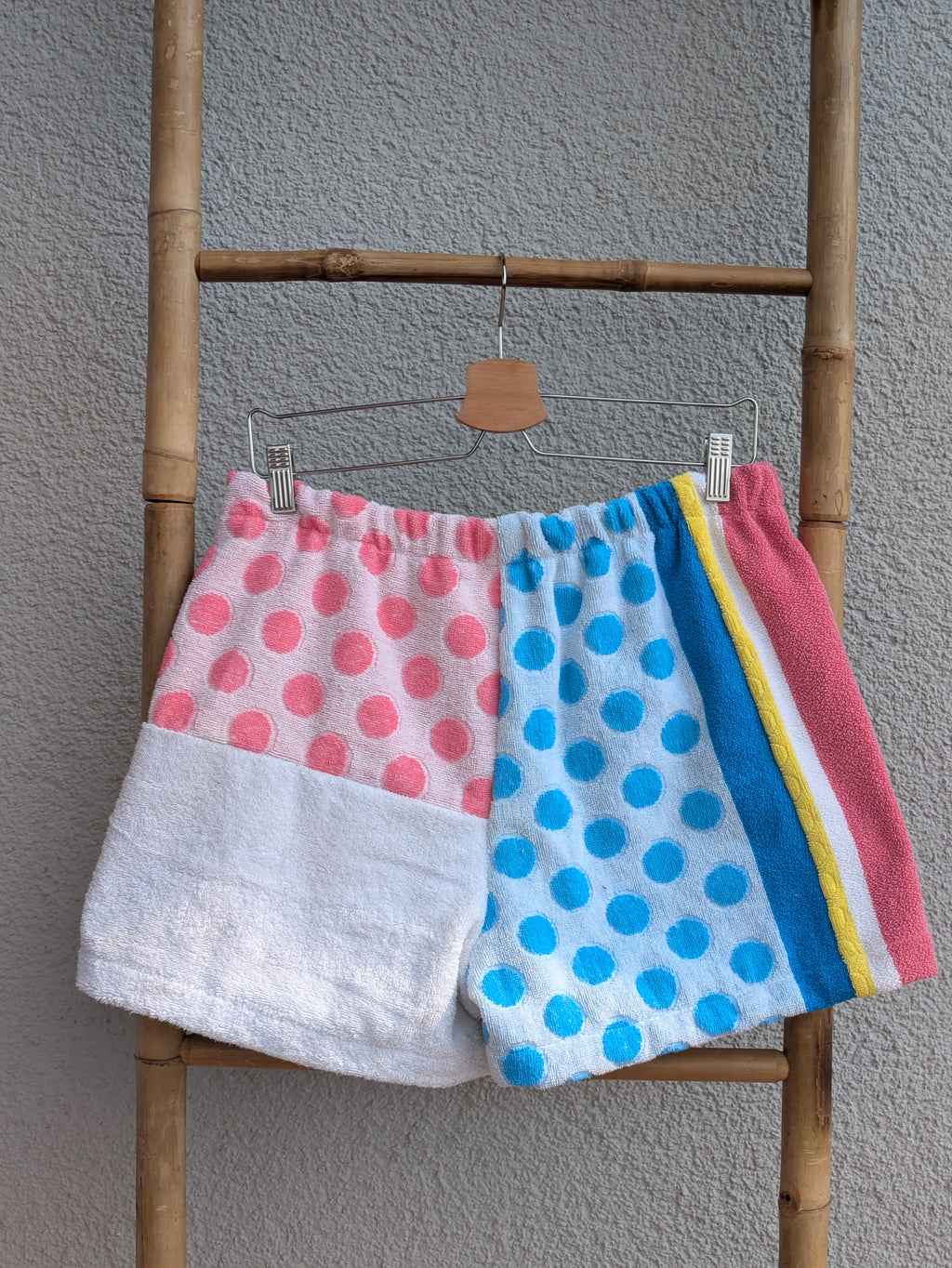 Rückansicht der Frottee Towel Shorts 'Retro Summer Circus ’78' von Old Wave. Zeigt das verspielte Patchwork-Design mit rosa Punkten, blauem Dot-Muster, weißem Hosenbein-Einsatz und bunten Streifen vor einer hellen Wand.