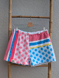 Vorderansicht der Frottee Towel Shorts 'Retro Summer Circus ’78' von Old Wave. Bunter Patchwork-Mix mit rosa und blauen Polka-Dot-Punkten, farbenfrohen Längs- und Querstreifen in Pink, Blau und Gelb sowie Old Wave Logo-Patch unten links vor einer Bambusleiter.