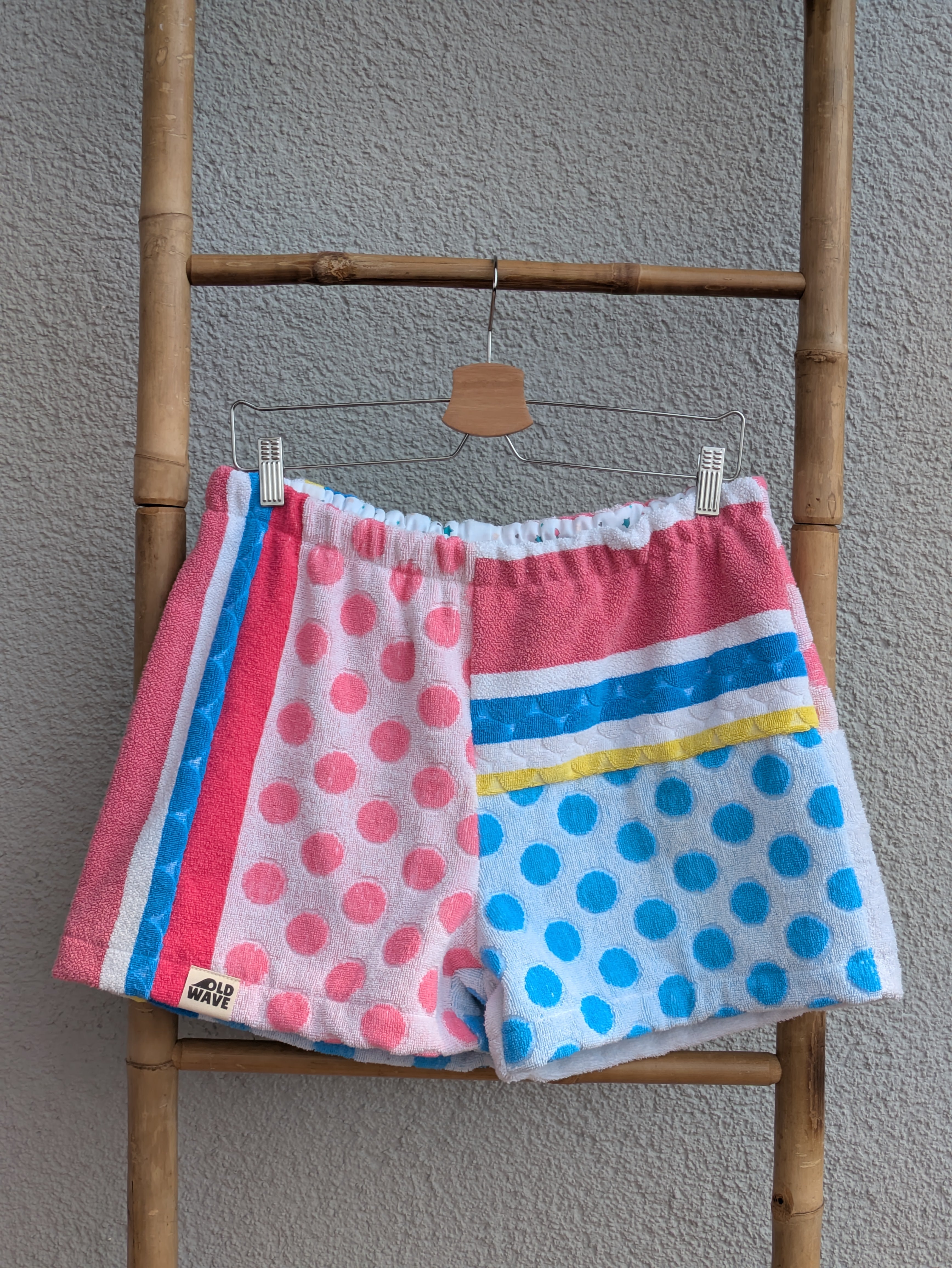 Vorderansicht der Frottee Towel Shorts 'Retro Summer Circus ’78' von Old Wave. Bunter Patchwork-Mix mit rosa und blauen Polka-Dot-Punkten, farbenfrohen Längs- und Querstreifen in Pink, Blau und Gelb sowie Old Wave Logo-Patch unten links vor einer Bambusleiter.