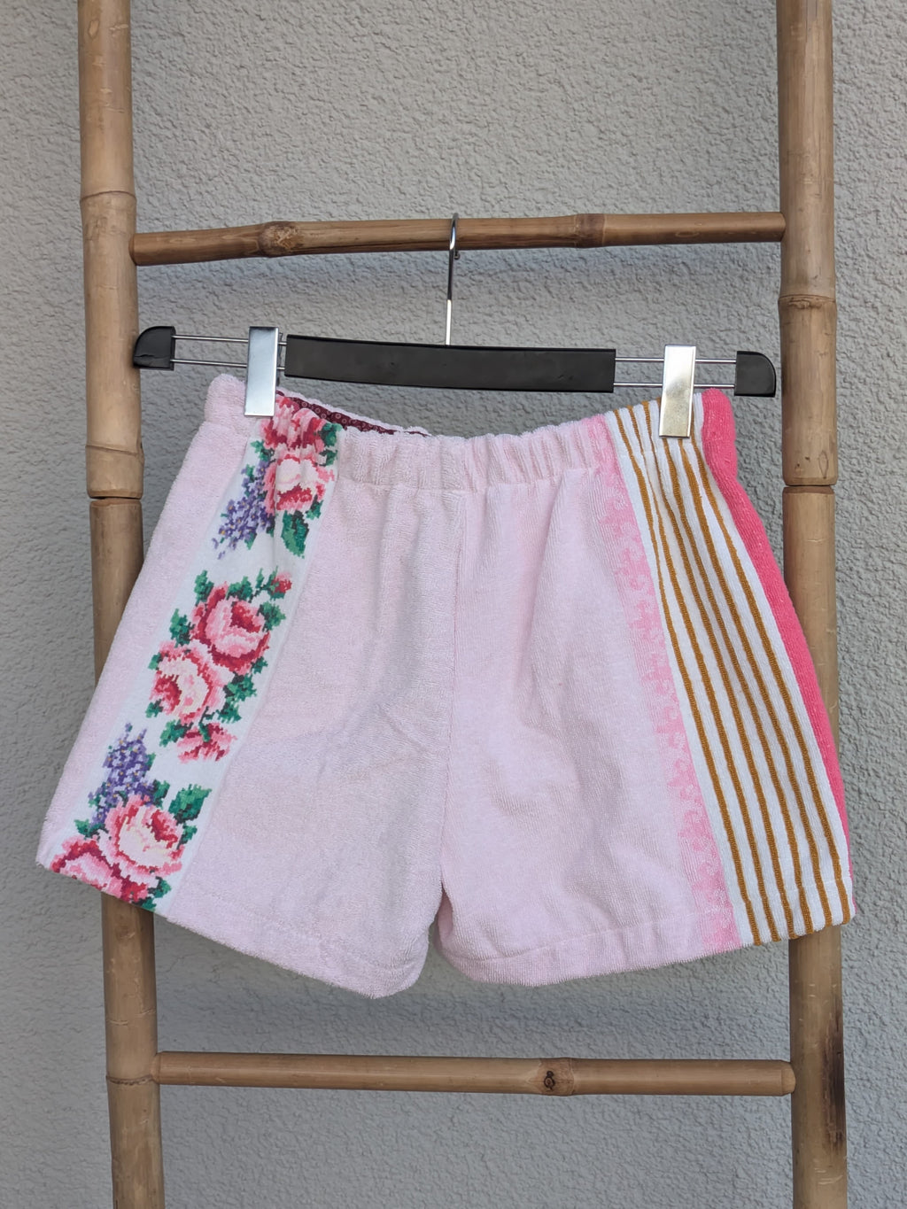 Rückansicht der Old Wave Towel Shorts 'Rose Blossom ’74'. Zeigt die rosafarbene Rückseite mit seitlichen Details: links das floral gemusterte Frottee, rechts ein beiger Streifeneinsatz mit pinker Umrandung. Die Shorts hängen an einer Bambusleiter.