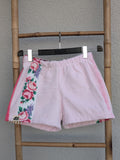 Vorderansicht der Frottee-Shorts 'Rose Blossom ’74' von Old Wave. Zartrosafarbene Shorts mit einem bunten Rosen-Musterstreifen auf der einen Seite und knallpinken Akzentpaspeln. Die Hose hängt an einem Kleiderbügel vor einer hellen Wand.