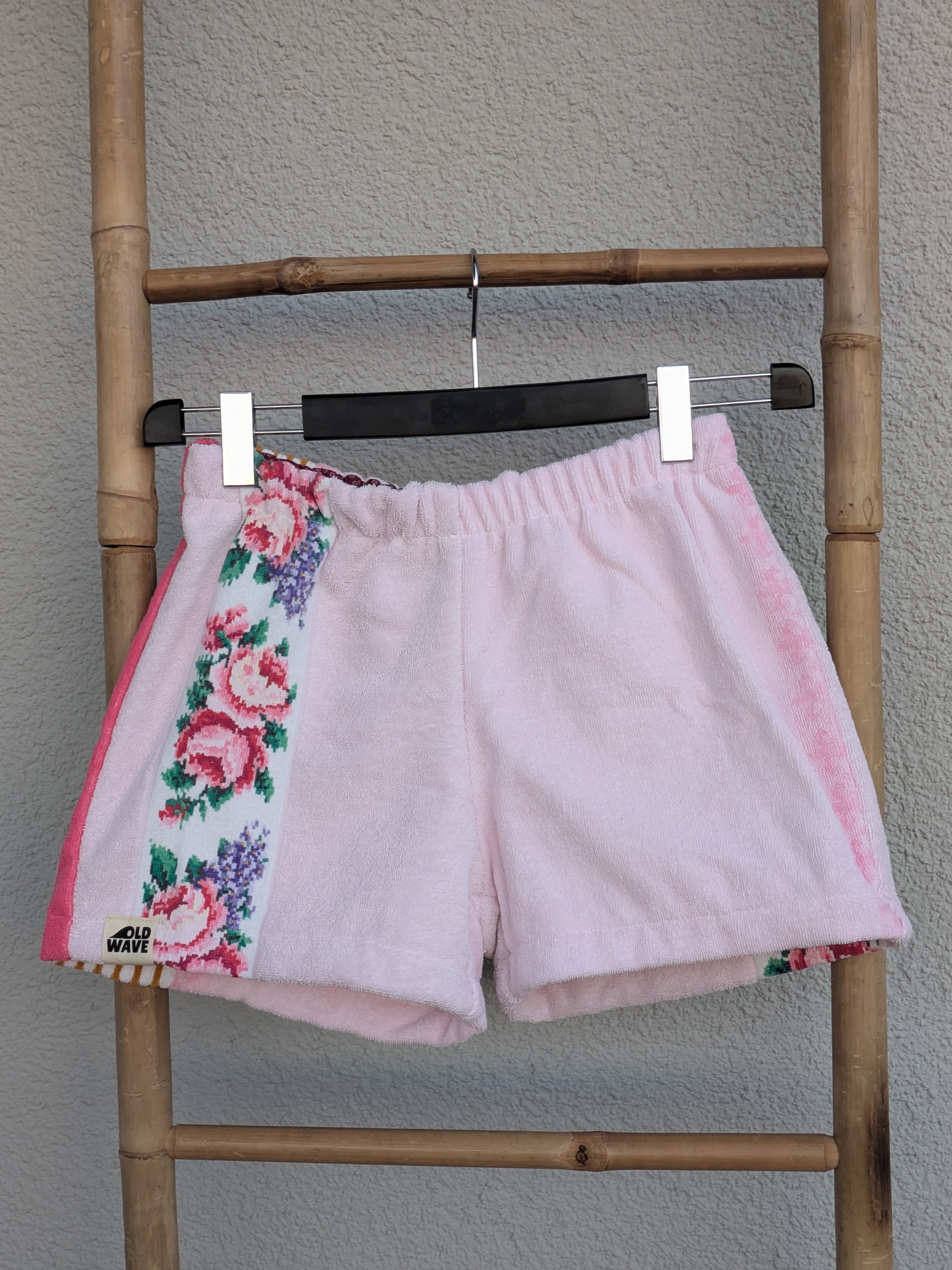 Vorderansicht der Frottee-Shorts 'Rose Blossom ’74' von Old Wave. Zartrosafarbene Shorts mit einem bunten Rosen-Musterstreifen auf der einen Seite und knallpinken Akzentpaspeln. Die Hose hängt an einem Kleiderbügel vor einer hellen Wand.
