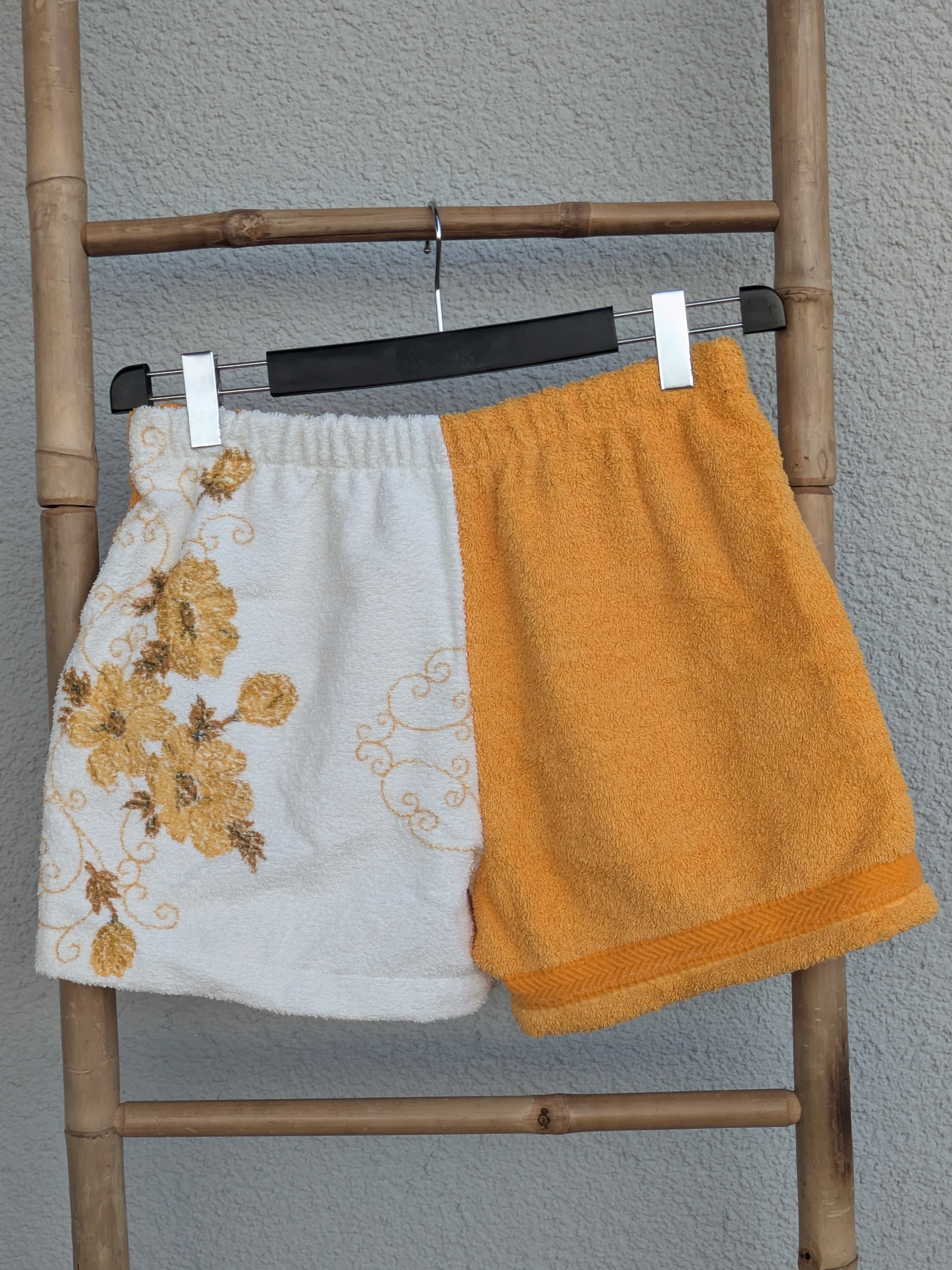 Rückansicht der zweifarbigen Old Wave Towel Shorts. Zeigt die Rückseite der weißen Hälfte mit dem Blumenmuster und die glatte, sonnengelbe rechte Hälfte. Die Hose hängt an einem Kleiderbügel vor einer strukturierten Wand.