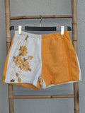 Nahaufnahme der Old Wave Frottee-Shorts im Retro-Look. Die linke Hälfte ist weiß mit aufgestickten senfgelben Retro-Blumen, die rechte Hälfte ist uni sonnengelb. Oben ein bequemer Gummibund, unten links das dezente Old Wave Logo-Patch. Die Hose hängt an einem Kleiderbügel vor einer hellgrauen Wand.