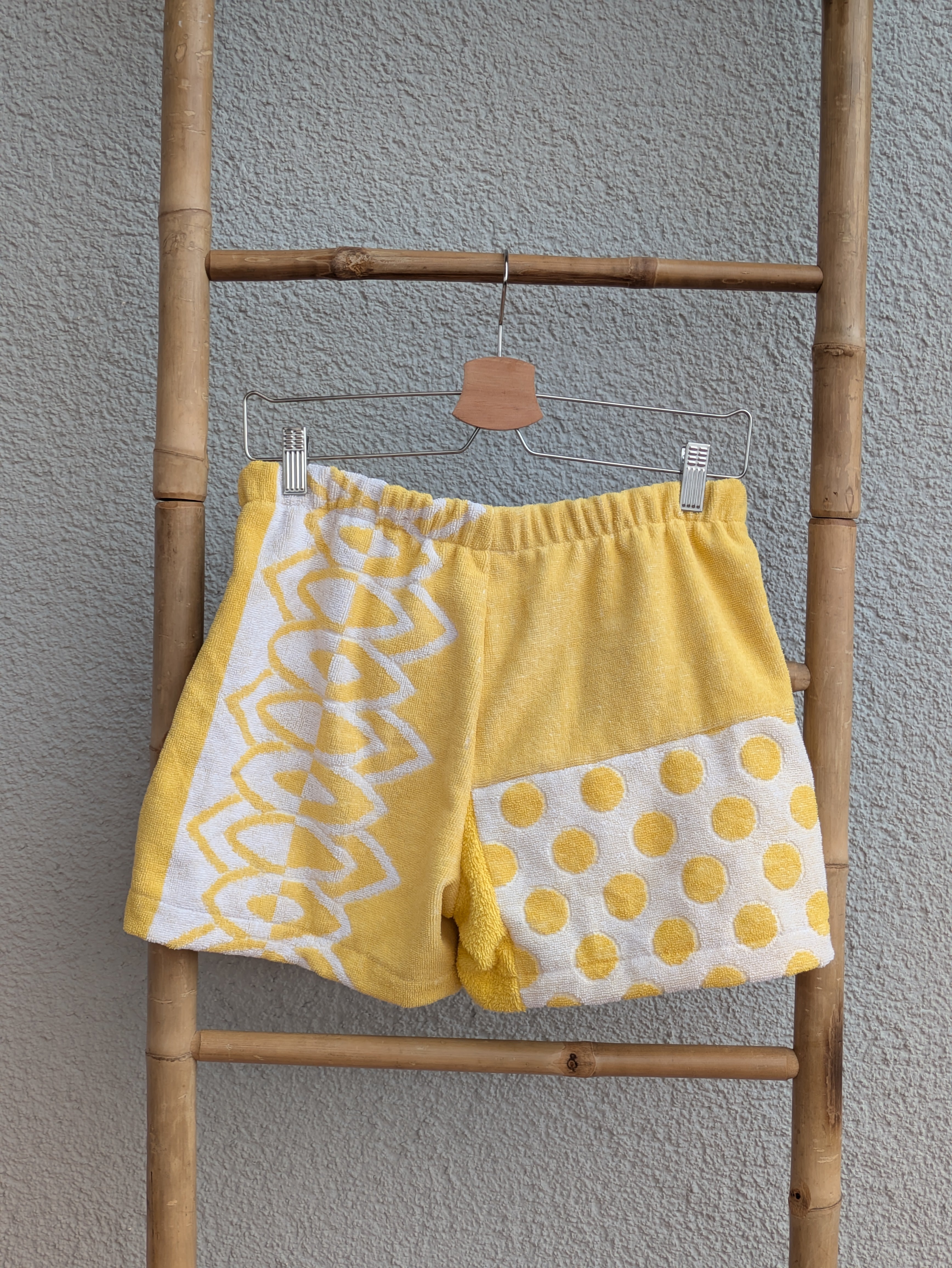 Rückansicht der Frottee Towel Shorts 'Sunny Heart Ornament ’77' von Old Wave. Zeigt das Patchwork-Design mit unifarbenem gelben Frottee oben rechts, gelb-weißem Polka-Dot-Punktemuster unten und Geometrie-Muster links vor einer hellen Wand.