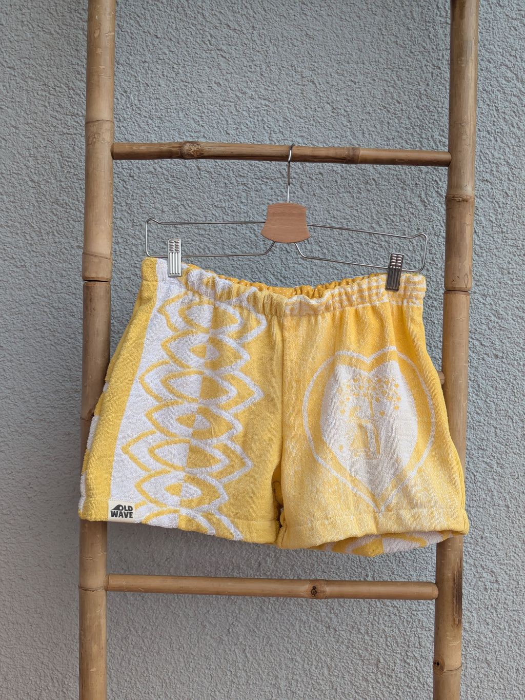 Vorderansicht der Frottee Towel Shorts 'Sunny Heart Ornament ’77' von Old Wave in Leuchtgelb und Weiß. Patchwork-Design mit großem eingewebten Herzmotiv rechts, geometrischem Bogen-Muster links und Logo-Patch unten links, präsentiert an einer Bambusleiter.