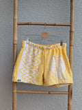 Vorderansicht der Frottee Towel Shorts 'Sunny Heart Ornament ’77' von Old Wave in Leuchtgelb und Weiß. Patchwork-Design mit großem eingewebten Herzmotiv rechts, geometrischem Bogen-Muster links und Logo-Patch unten links, präsentiert an einer Bambusleiter.