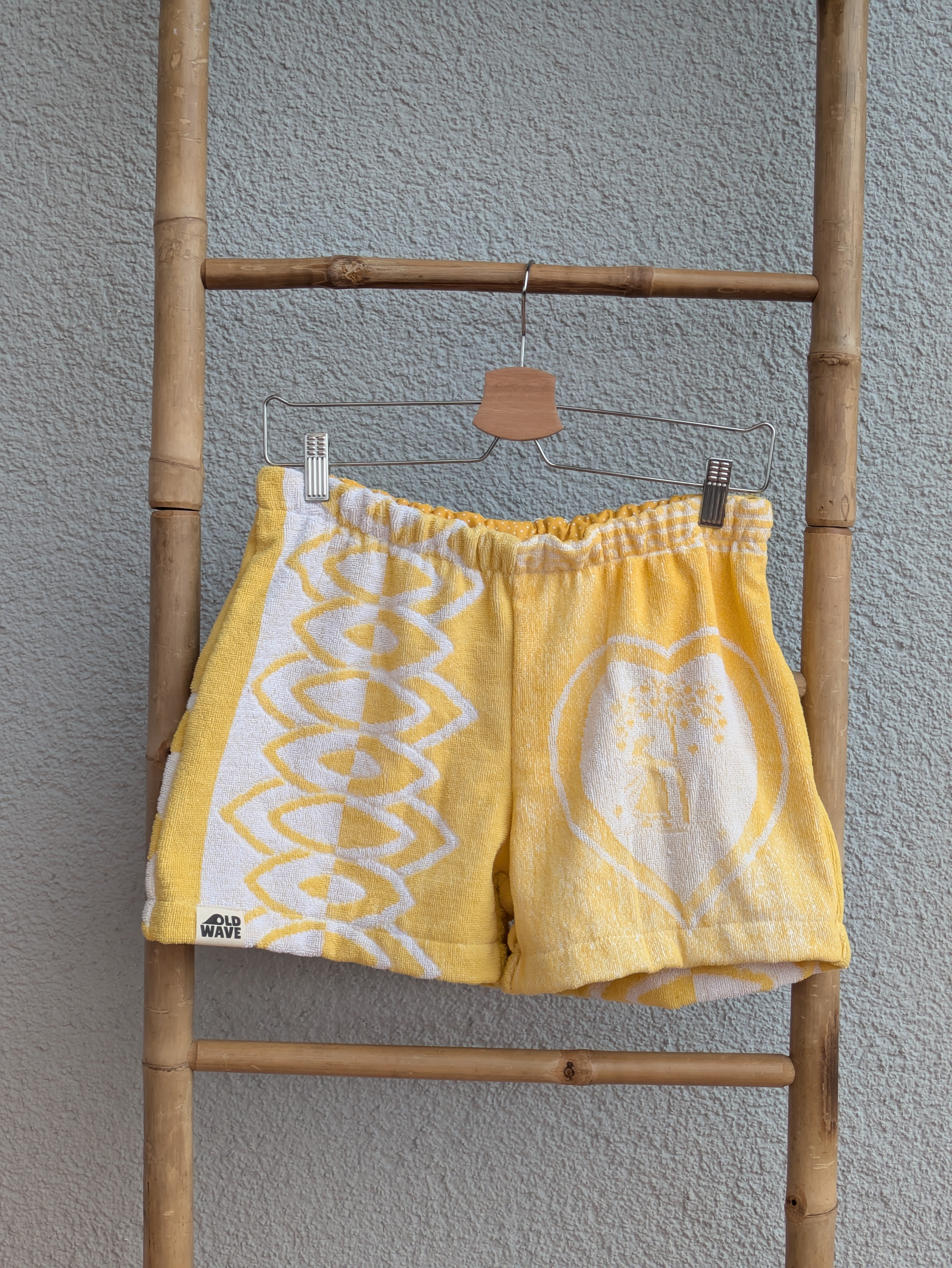Vorderansicht der Frottee Towel Shorts 'Sunny Heart Ornament ’77' von Old Wave in Leuchtgelb und Weiß. Patchwork-Design mit großem eingewebten Herzmotiv rechts, geometrischem Bogen-Muster links und Logo-Patch unten links, präsentiert an einer Bambusleiter.