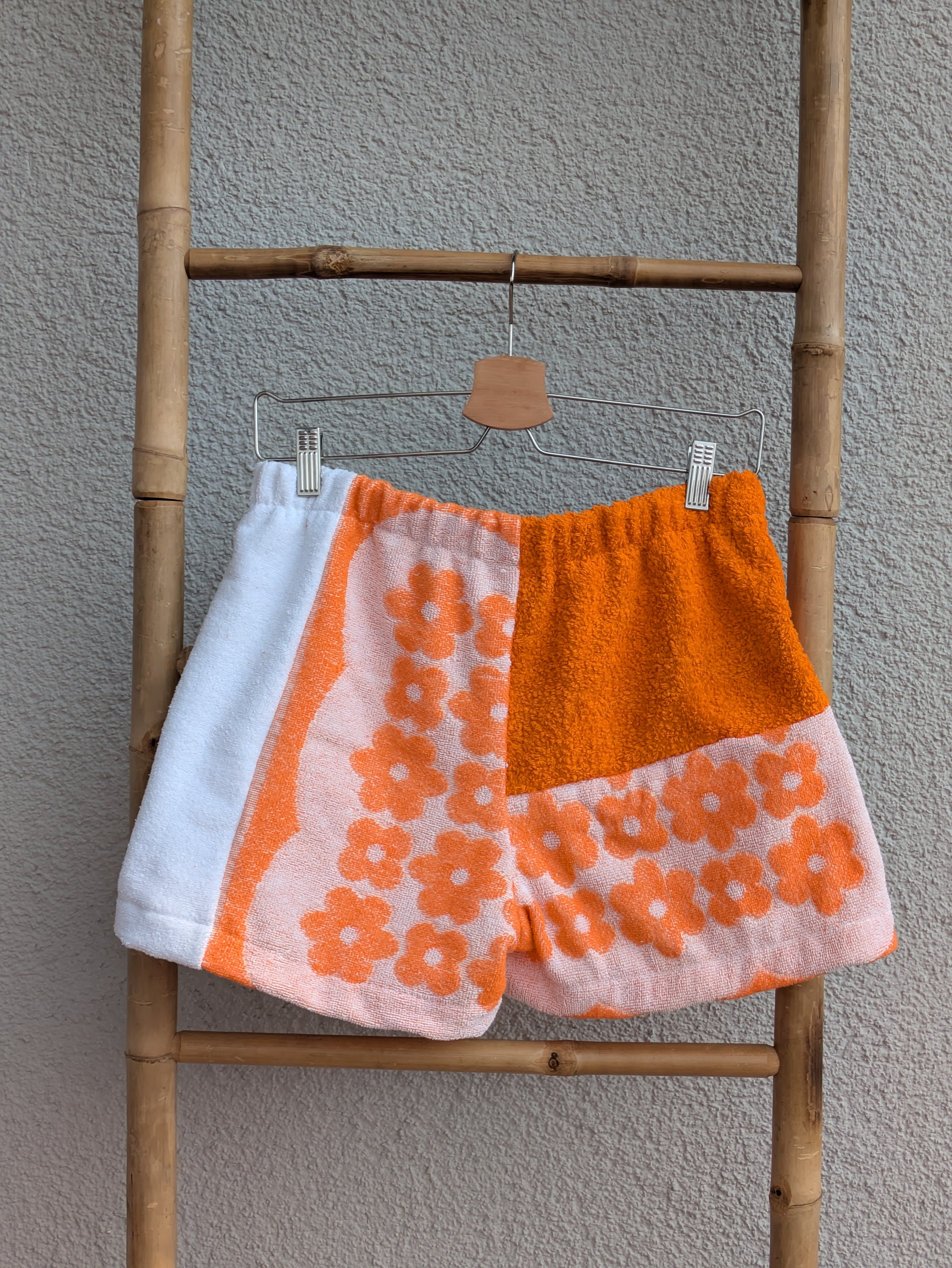 Rückansicht der Frottee Towel Shorts 'Sunset Floral Patch ’75' von Old Wave. Zeigt das Patchwork-Design mit unifarbenem Orange oben rechts und floralem 70s-Muster sowie weißem Seiteneinsatz vor einer hellen Wand.