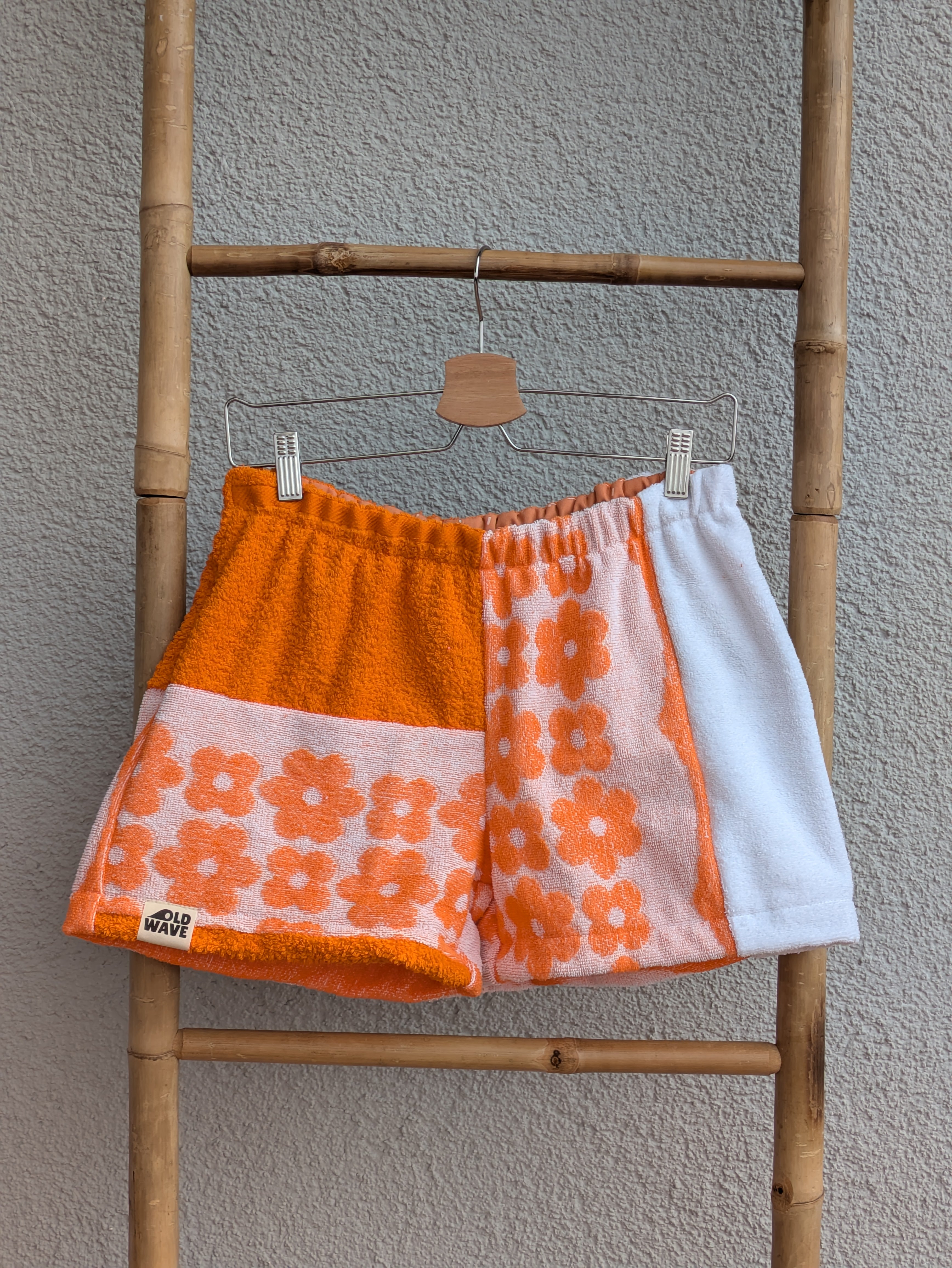 Vorderansicht der Frottee Towel Shorts 'Sunset Floral Patch ’75' von Old Wave. Patchwork-Design mit unifarbenem orangefarbenem Hosenbein und floralem Retro-Muster, weißem Seiteneinsatz sowie Old Wave Logo-Patch unten links vor einer Bambusleiter.