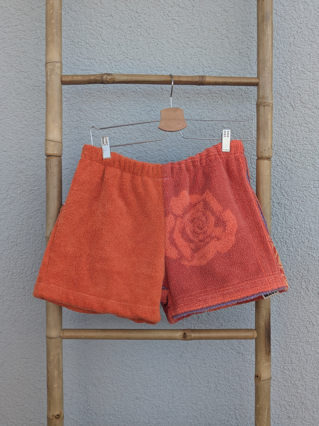 Rückansicht der Frottee Towel Shorts 'Terracotta Floral ’72' von Old Wave. Zeigt das zweigeteilte Two-Tone-Design mit dem großen eingewebten Rosenmotiv auf der linken Gesäßhälfte vor einer hellen Wand.