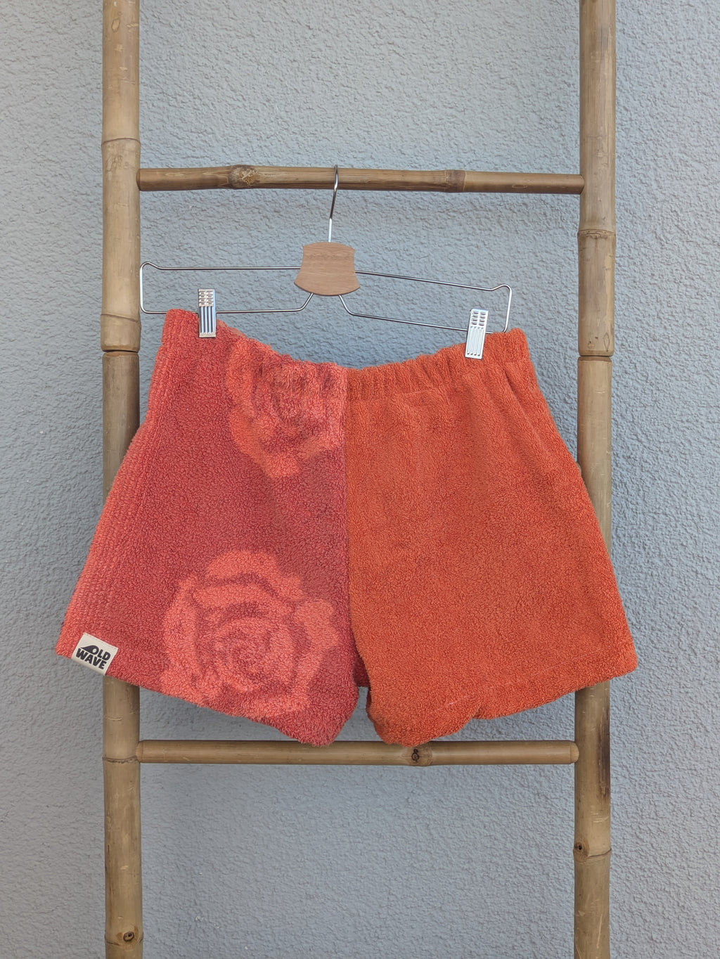 Vorderansicht der Frottee Towel Shorts 'Terracotta Floral ’72' von Old Wave in Korallenrot und Terracotta. Zweigeteiltes Patchwork-Design mit großem Rosenmotiv auf der rechten Beinseite und Old Wave Logo-Patch unten links, präsentiert an einer Bambusleiter.