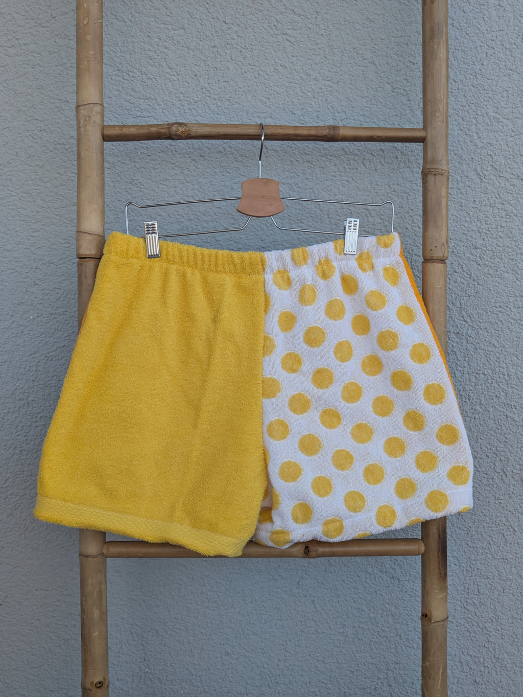 Rückansicht der Frottee Towel Shorts 'Yellow Polka ’76' von Old Wave. Zeigt das zweigeteilte Half-and-Half-Design in unifarbenem Sonnengelb und weißem Frottee mit großen gelben Punkte-Muster vor einer hellen Wand.