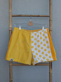 Vorderansicht der Frottee Towel Shorts 'Yellow Polka ’76' von Old Wave in Sonnengelb und Weiß. Zweigeteiltes Two-Tone-Design mit unifarbener gelber Hosenhälfte, gelb-weiß gepunktetem Polka-Dot-Muster und gelbem Seiteneinsatz, präsentiert an einer Bambusleiter.