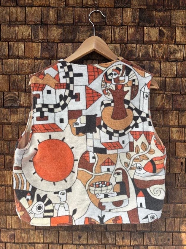 Rückansicht der Frottee-Weste 'Clementine' von Old Wave. Zeigt das durchgehende, künstlerische 70s-Retromuster mit Sonnensymbolen und abstrakten Formen in Orange, Braun und Schwarz vor einer Backsteinwand.