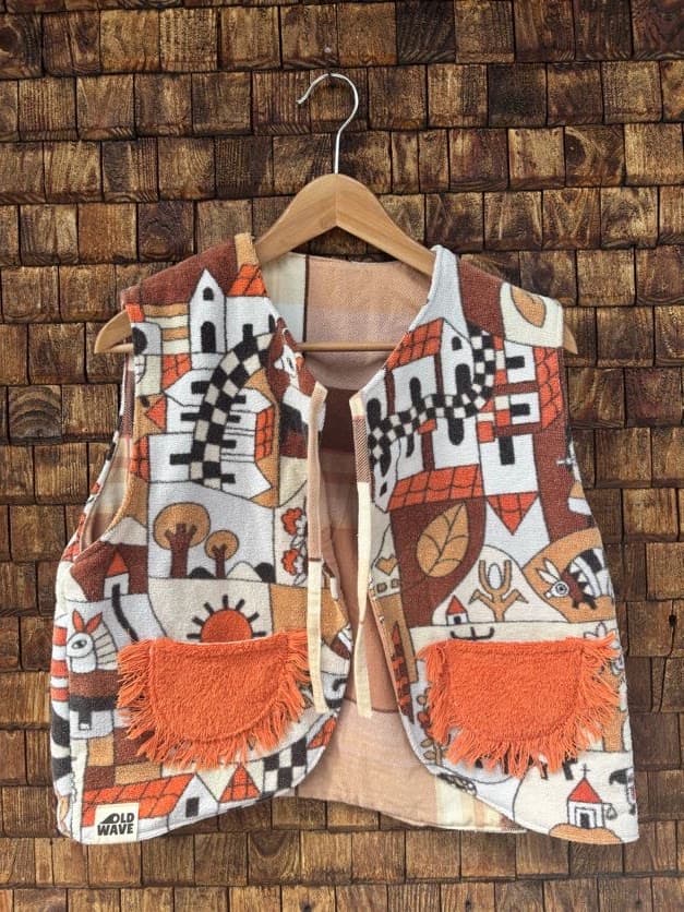 Vorderansicht der wendbaren Frottee-Weste 'Clementine' von Old Wave. Ärmellose Weste mit auffälligem 70s-Retro-Print in Orange, Braun, Schwarz und Cremeweiß sowie zwei orangefarbenen aufgesetzten Taschen vor einer Backsteinwand.