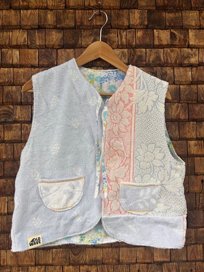 Vorderansicht der wendbaren Frottee-Weste 'Cloud Reversible ’72' von Old Wave. Ärmellose Weste in Wolkenblau und Weiß mit filigranen Ornament-Mustern, rosafarbener Zierborte und zwei aufgesetzten Taschen vor einer Backsteinwand.