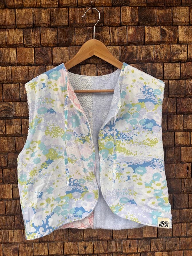 Vorderansicht der gewendeten Frottee-Weste 'Cloud Reversible ’72' von Old Wave. Zweite Trageoption mit bunter Pastell-Musterung in Hellblau, Gelb und Grün auf weißem Frottee vor einer Backsteinwand.