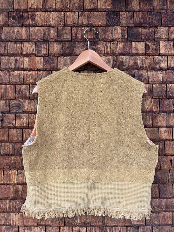 Rückansicht der Frottee-Weste 'Dune' von Old Wave. Zeigt die Rückseite in warmem Sandbeige mit Fransenkante am unteren Saum vor einer Backsteinwand.
