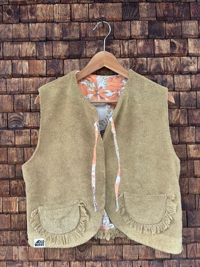 Vorderansicht der wendbaren Frottee-Weste 'Dune' von Old Wave. Ärmellose Weste in unifarbenem Sandbeige mit dezent eingewebtem Ornamentmuster, zwei aufgesetzten Taschen und Fransen-Abschluss am Saum vor einer Backsteinwand.