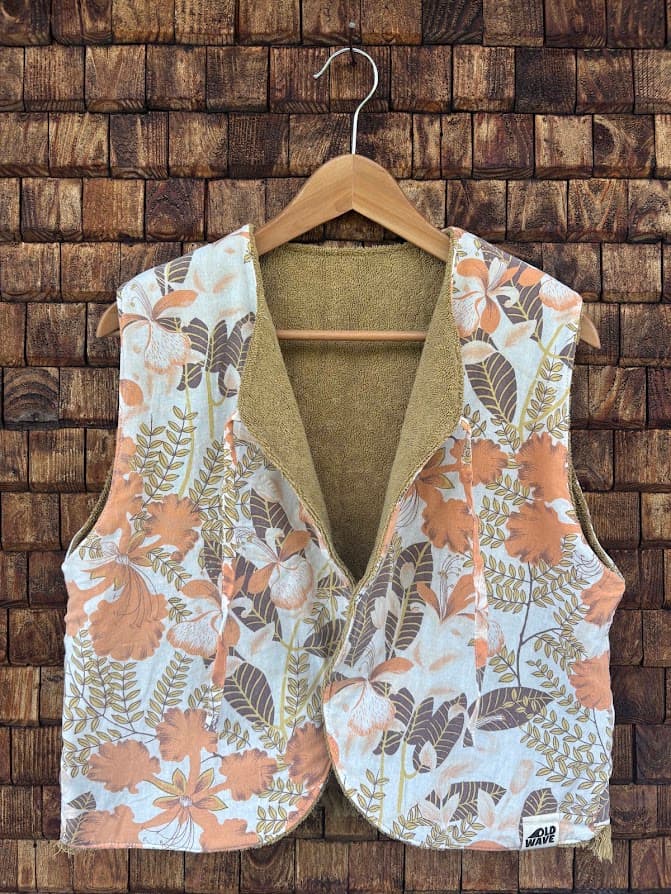 Vorderansicht der gewendeten Frottee-Weste 'Dune' von Old Wave. Zweite Trageoption mit einem großflächigen 70s-Blumenmuster in Terracotta, Orange und Beige vor einer Backsteinwand.
