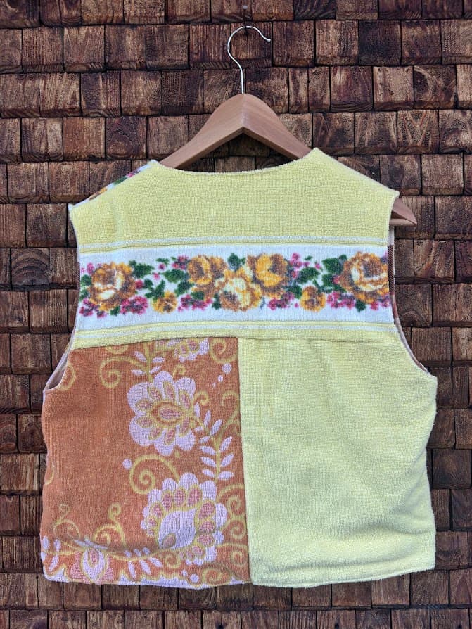 Rückansicht der Frottee-Weste 'Honey' von Old Wave. Zeigt das Patchwork-Design mit sonnengelber Hälfe, orangenem Retro-Blumenmuster und quer verlaufender Blumenborte im Rücken vor einer Backsteinwand.