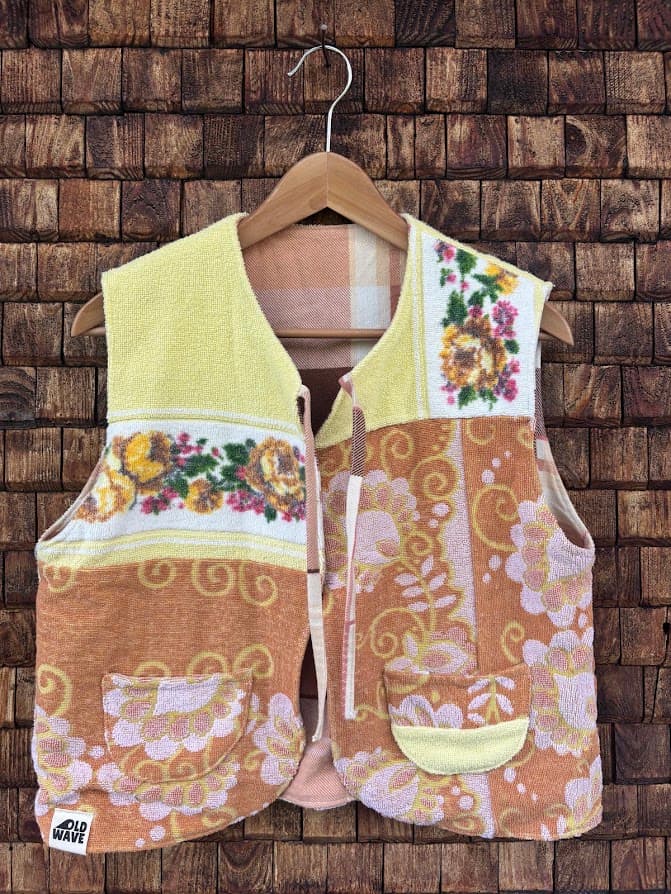 Vorderansicht der wendbaren Frottee-Weste 'Honey' von Old Wave. Ärmellose Patchwork-Weste in Sonnengelb, Senfgelb und Orange mit floraler Borte und Ornamentmuster sowie einer kleinen gelben Tasche vor einer Backsteinwand.
