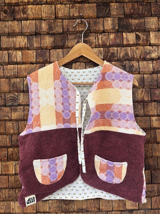 Vorderansicht der wendbaren Frottee-Weste 'Plum' von Old Wave. Ärmellose Weste in tiefem Pflaumenrot/Bordeaux mit Retromuster-Borte im Brustbereich in Orange, Violett und Gelb sowie zwei Taschen mit passendem Muster vor einer Backsteinwand.