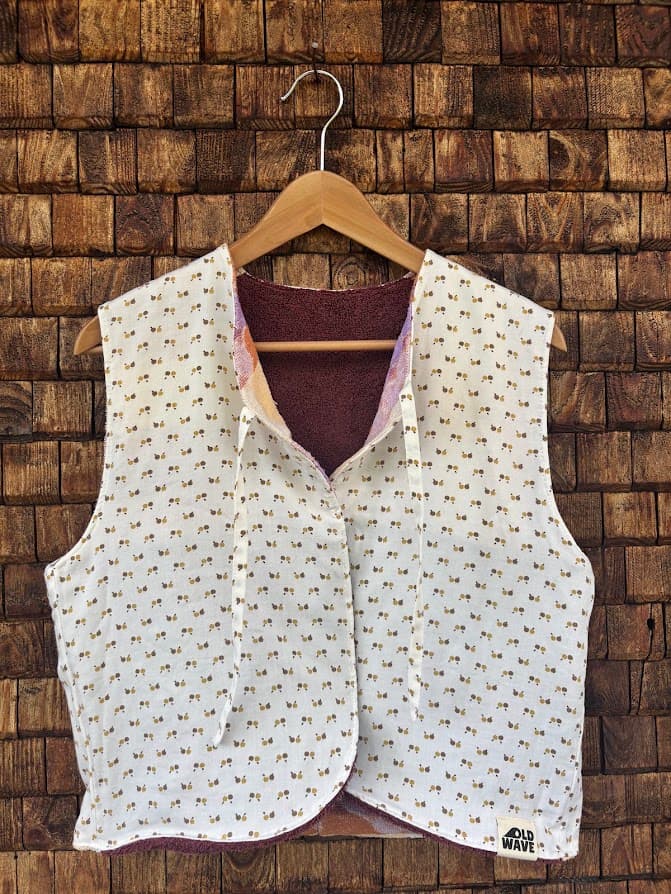 Vorderansicht der gewendeten Frottee-Weste 'Plum' von Old Wave. Zweite Trageoption in hellem Off-White / Cremeweiß mit dezentem, feinem Pünktchen- und Rautenmuster vor einer Backsteinwand.