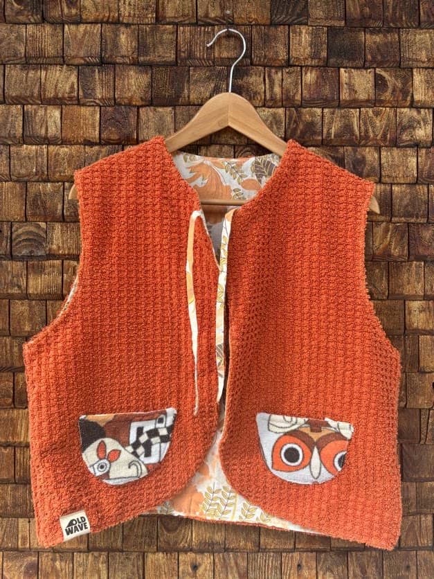 Vorderansicht der wendbaren Frottee-Weste 'Rust' von Old Wave. Ärmellose Weste in leuchtendem Rostorange mit zwei aufgesetzten Taschen im Retromuster mit Eulen-Motiven vor einer Backsteinwand.