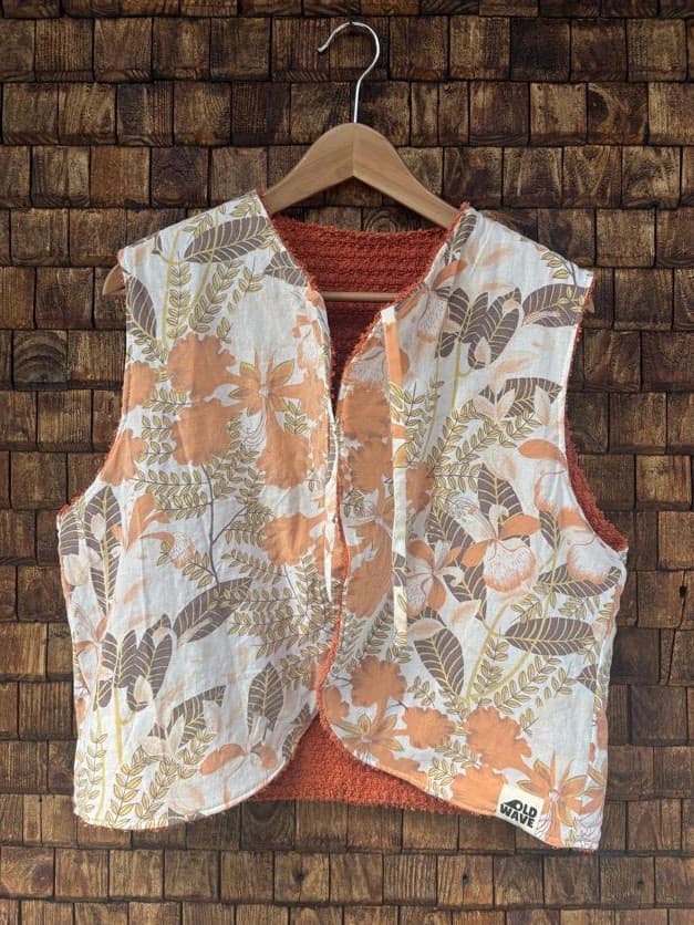 Vorderansicht der gewendeten Frottee-Weste 'Rust' von Old Wave. Zweite Trageoption mit einem großflächigen 70s-Blumenmuster in Apricot, Orange und Braun vor einer Backsteinwand.