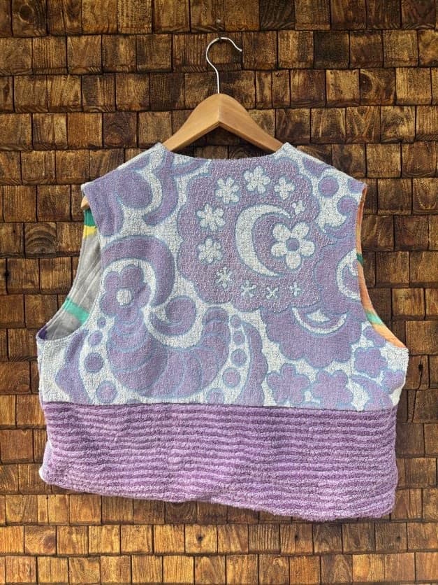 Rückansicht der Frottee-Weste 'Violet' von Old Wave. Zeigt das durchgehende, farbenfrohe Retro-Blumenmuster in knalligen Farben auf der Rückseite vor einer Backsteinwand.