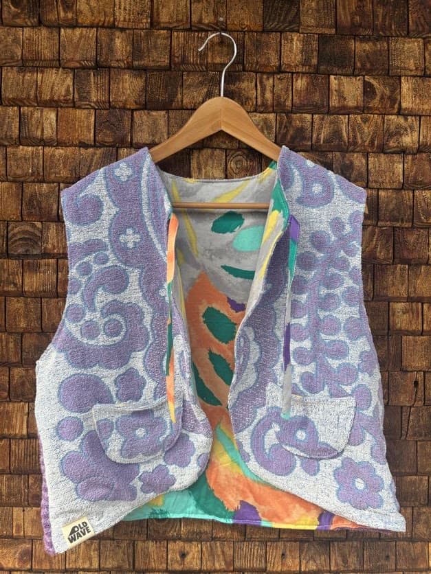 Vorderansicht der wendbaren Frottee-Weste 'Violet' von Old Wave. Ärmellose Weste mit lebendigem Psychedelic-Print in Violett, Türkis, Orange, Gelb und Grau vor einer Backsteinwand.