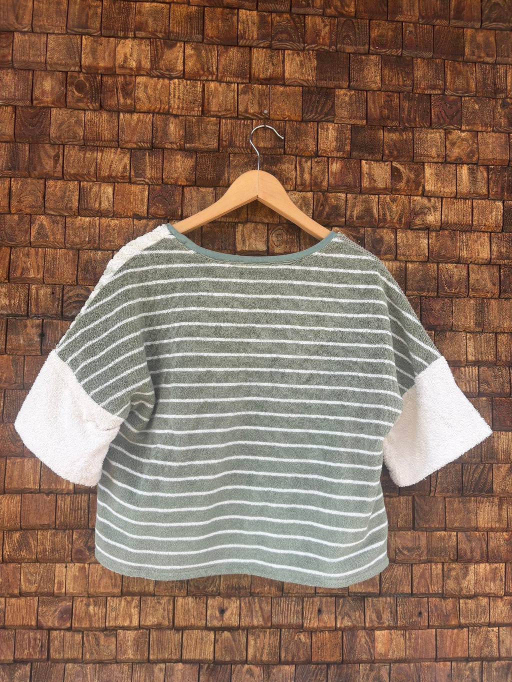 Rückansicht des Upcycling-Frotteeshirts „Olive Wave ‘74“ von Old Wave auf einem Kleiderbügel vor einer Holzwand. Die Rückseite des lässig geschnittenen Oberteils präsentiert ein durchgehendes, horizontales Streifenmuster in Olivgrün und Weiß. Dieses bildet einen harmonischen Kontrast zu den unifarbenen weißen Ärmeln.