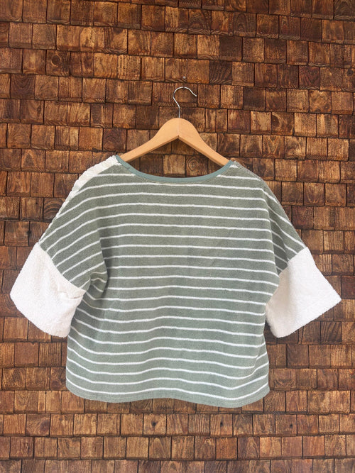 Rückansicht des Upcycling-Frotteeshirts „Olive Wave ‘74“ von Old Wave auf einem Kleiderbügel vor einer Holzwand. Die Rückseite des lässig geschnittenen Oberteils präsentiert ein durchgehendes, horizontales Streifenmuster in Olivgrün und Weiß. Dieses bildet einen harmonischen Kontrast zu den unifarbenen weißen Ärmeln.