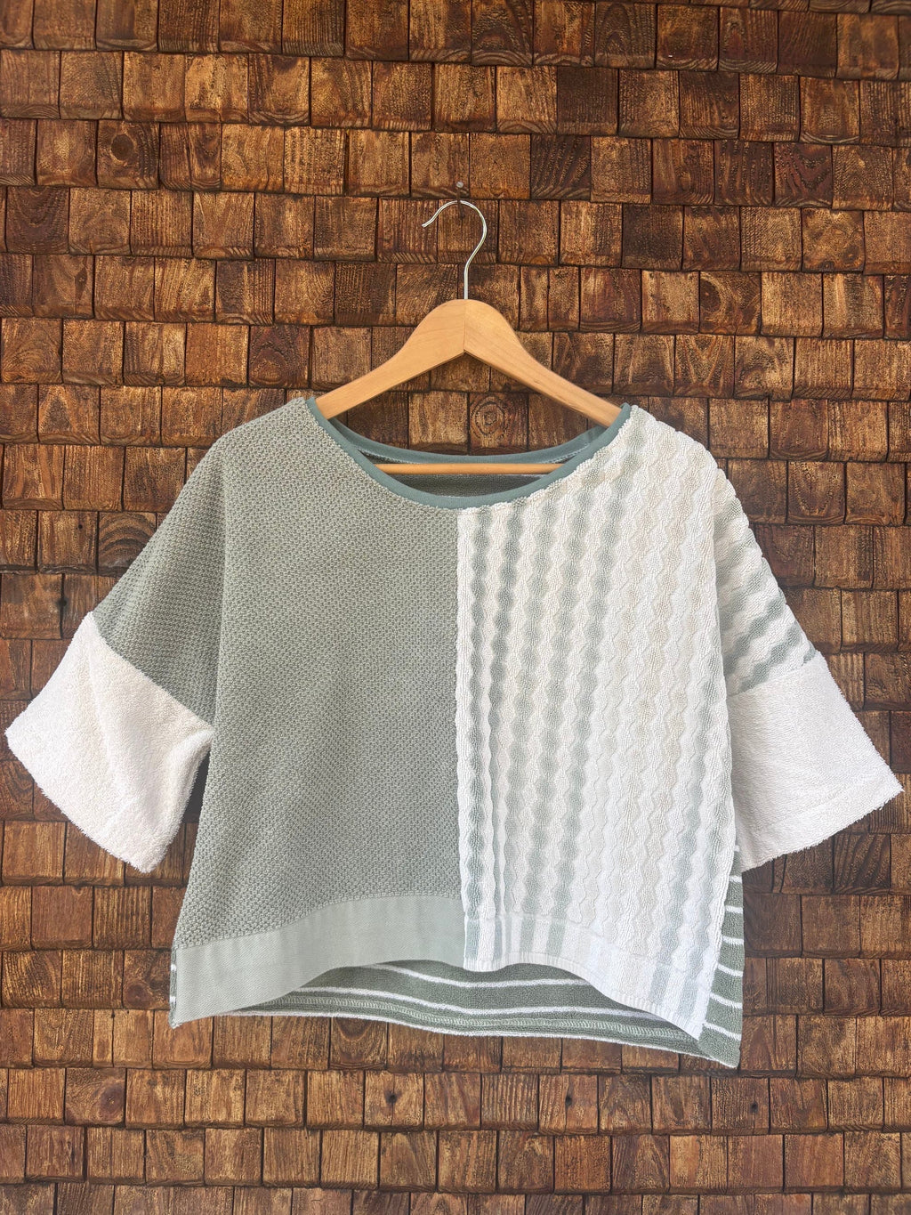 Vorderansicht des Upcycling-Frotteeshirts „Olive Wave ‘74“ von Old Wave auf einem Kleiderbügel vor einer rustikalen Holzwand. Das lockere Boxy-Shirt besticht durch ein zweigeteiltes Front-Design: Die linke Seite ist in schlichtem Salbei- bzw. Olivgrün gestaltet, während die rechte Seite aus weißem Vintage-Frottee mit einer vertikalen, reliefartig gewebten Musterstruktur besteht. Die weiten Ärmel sind in cleanem Weiß abgesetzt.
