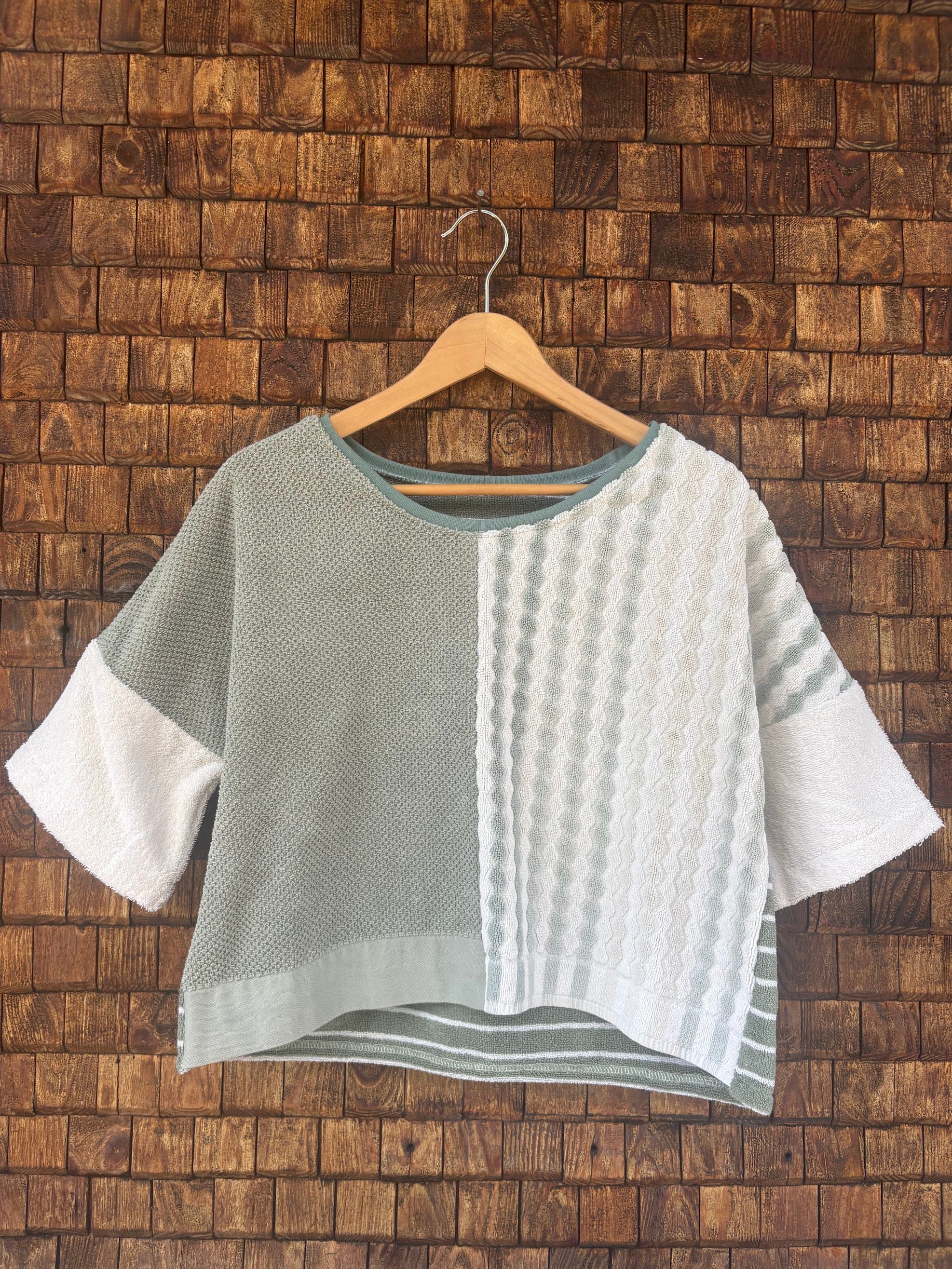 Vorderansicht des Upcycling-Frotteeshirts „Olive Wave ‘74“ von Old Wave auf einem Kleiderbügel vor einer rustikalen Holzwand. Das lockere Boxy-Shirt besticht durch ein zweigeteiltes Front-Design: Die linke Seite ist in schlichtem Salbei- bzw. Olivgrün gestaltet, während die rechte Seite aus weißem Vintage-Frottee mit einer vertikalen, reliefartig gewebten Musterstruktur besteht. Die weiten Ärmel sind in cleanem Weiß abgesetzt.