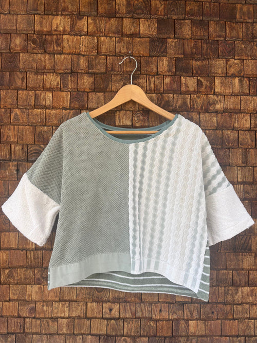 Vorderansicht des Upcycling-Frotteeshirts „Olive Wave ‘74“ von Old Wave auf einem Kleiderbügel vor einer rustikalen Holzwand. Das lockere Boxy-Shirt besticht durch ein zweigeteiltes Front-Design: Die linke Seite ist in schlichtem Salbei- bzw. Olivgrün gestaltet, während die rechte Seite aus weißem Vintage-Frottee mit einer vertikalen, reliefartig gewebten Musterstruktur besteht. Die weiten Ärmel sind in cleanem Weiß abgesetzt.